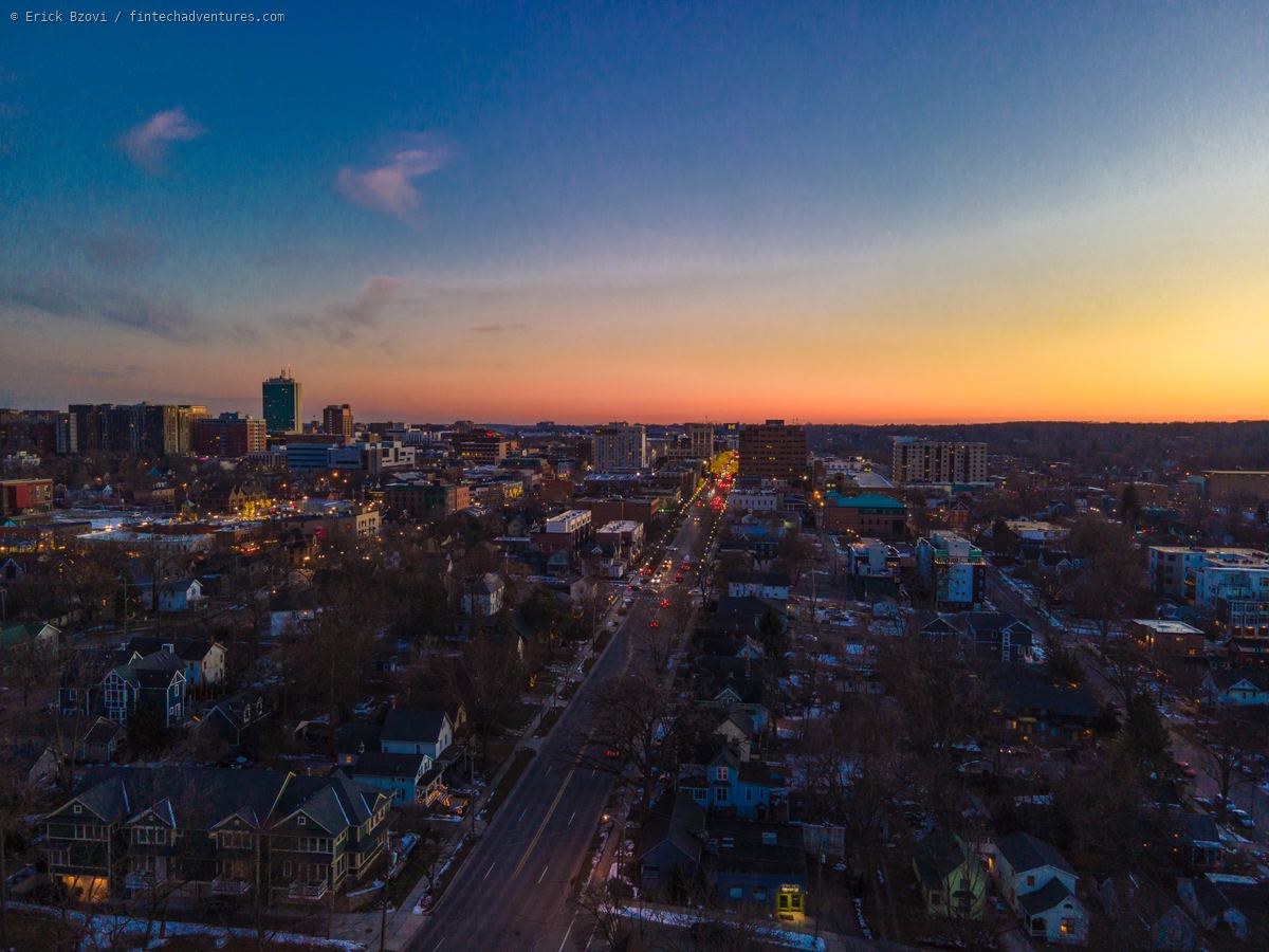 Aerial view at sunset over downtown Ann Arbor, Michigan, looking along a main road toward mid rise buildings, with bare winter trees, patchy snow and lit windows across the surrounding neighborhood