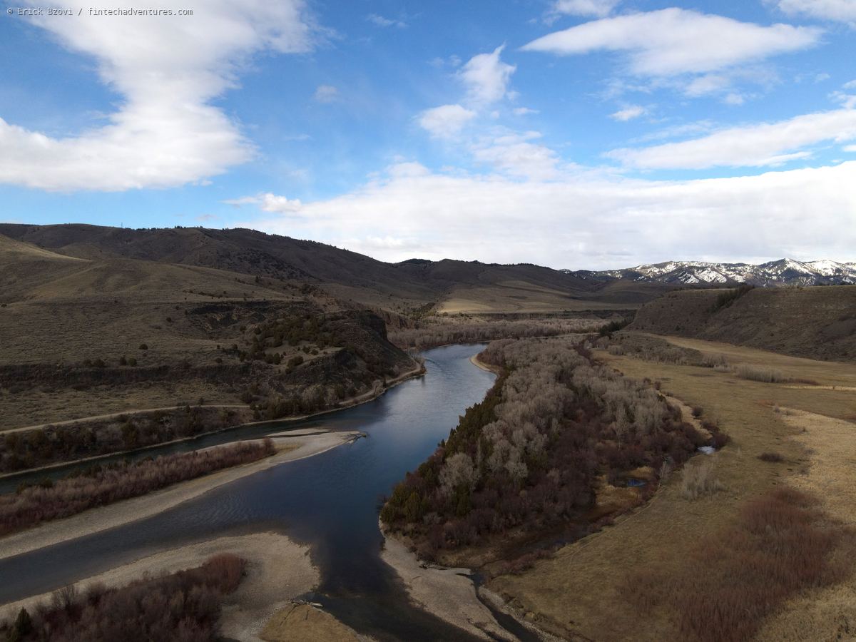 Aerial drone view of a river winding between gravel bars and bare cottonwoods through sage hills, snowcapped peaks in the distance, Idaho