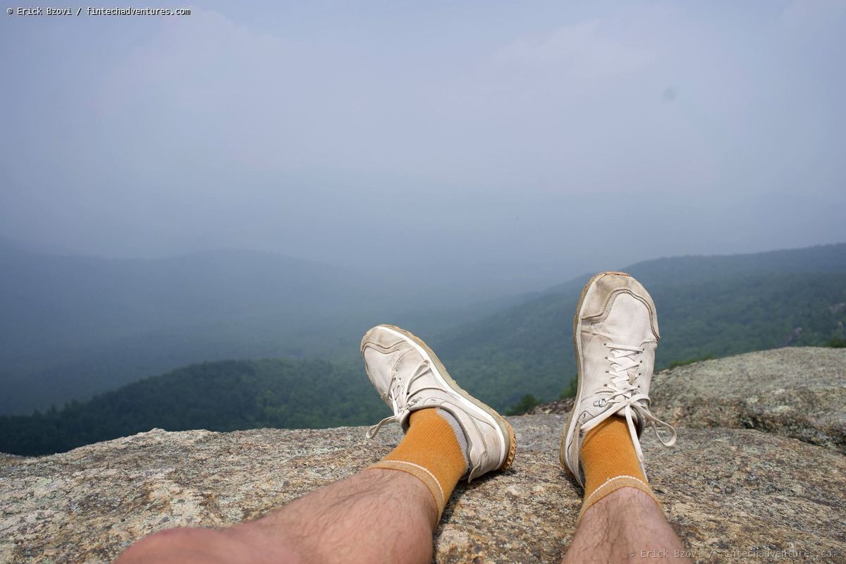 Hiking shoes over a rocky summit ledge with misty forested mountain ridges below